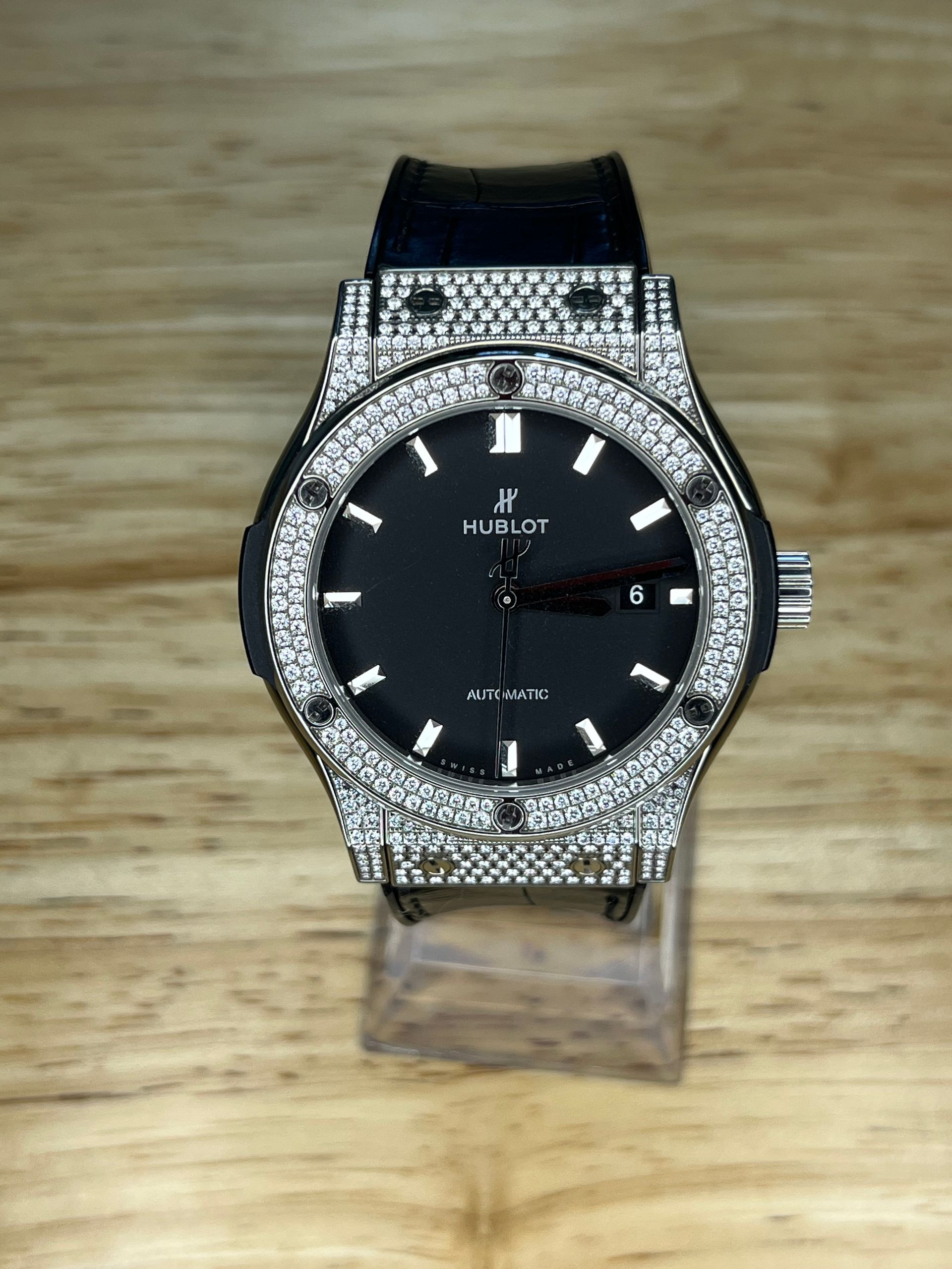 Diamond-encrusted Hublot watch with a black face, displayed on a clear stand against a wood background.