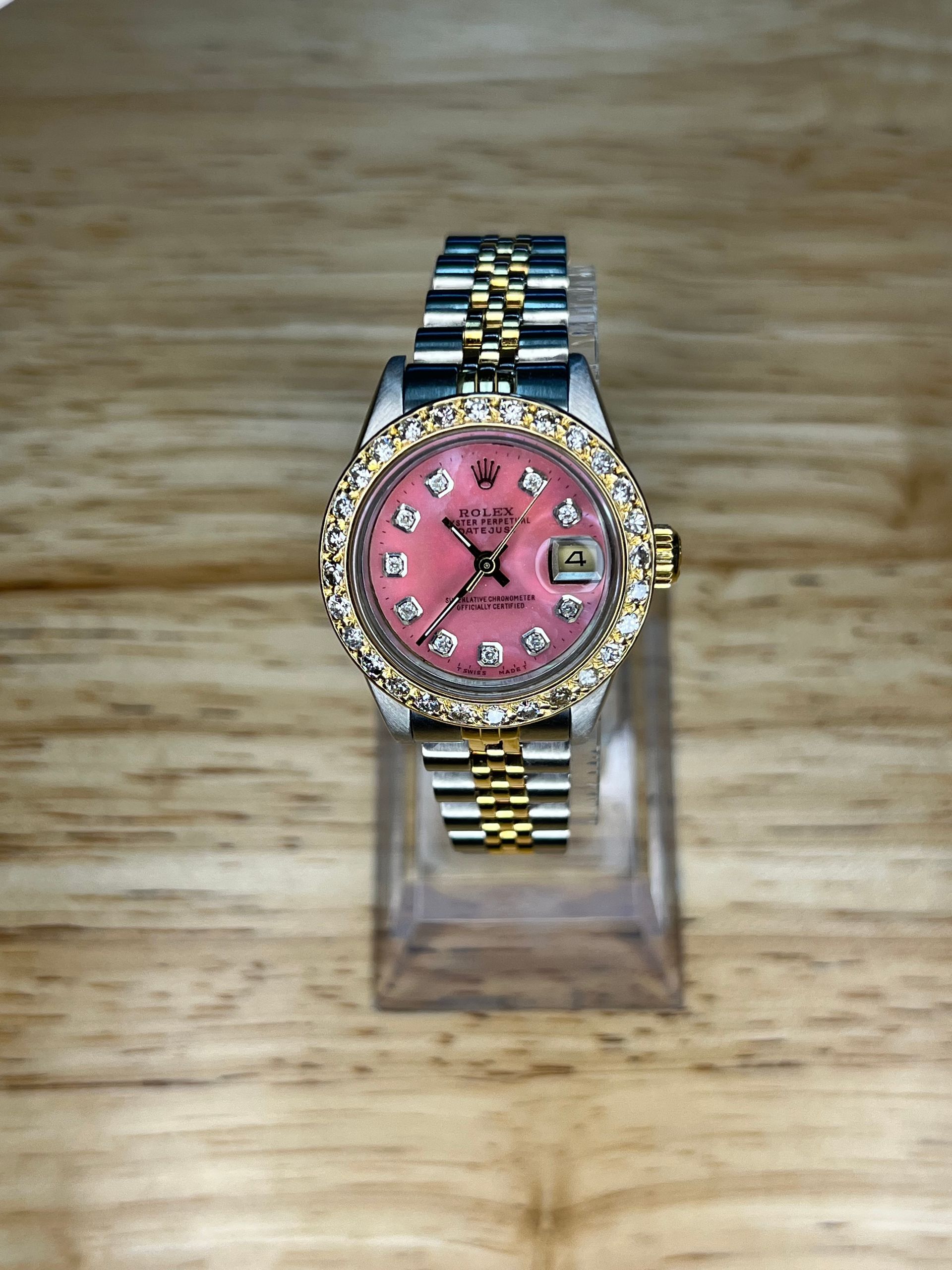 Rolex Datejust watch with a pink face and diamond-encrusted bezel, on a wooden surface.