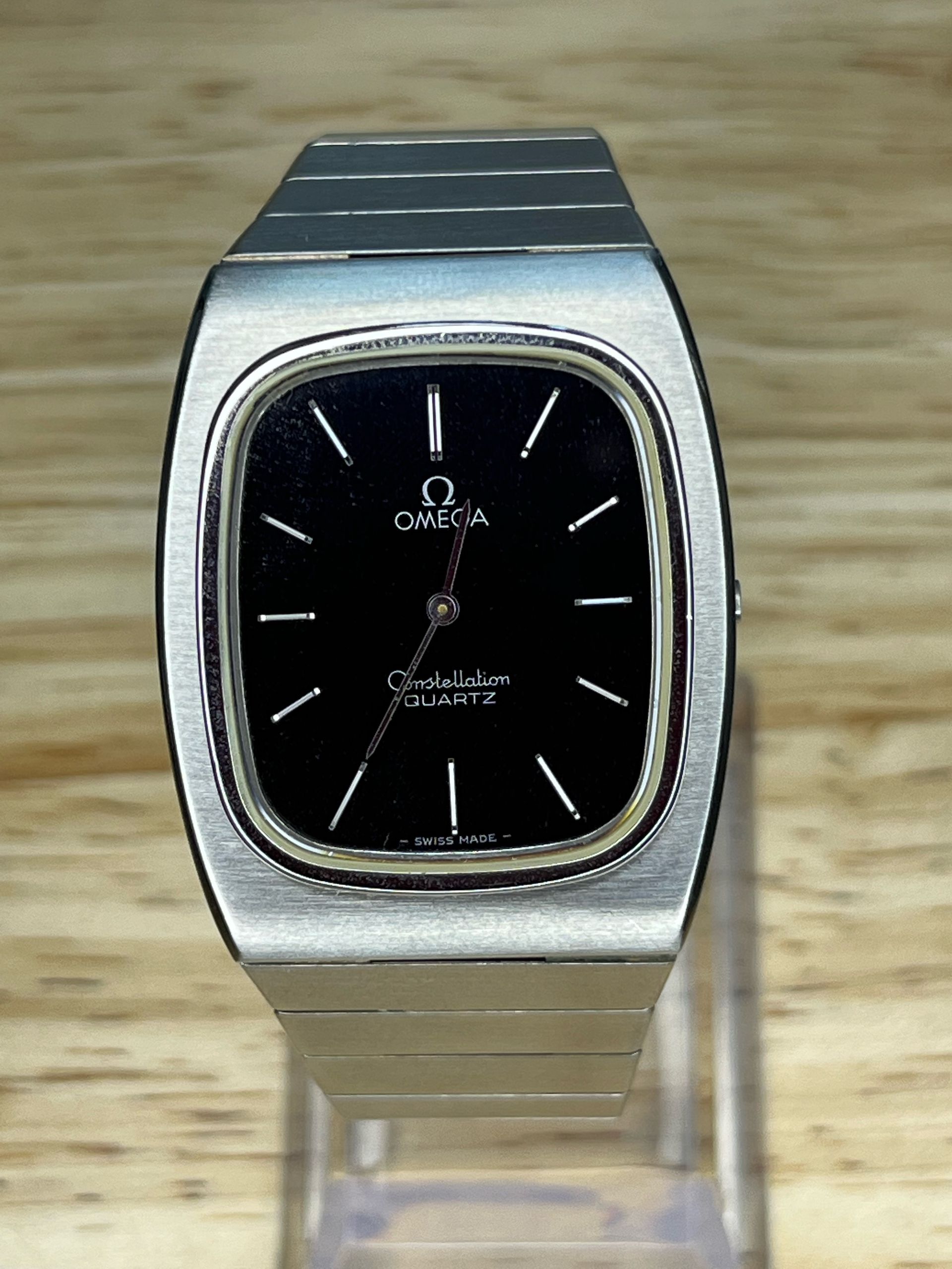 Silver Omega Quartz watch with a black dial, set against a light-colored wooden background.