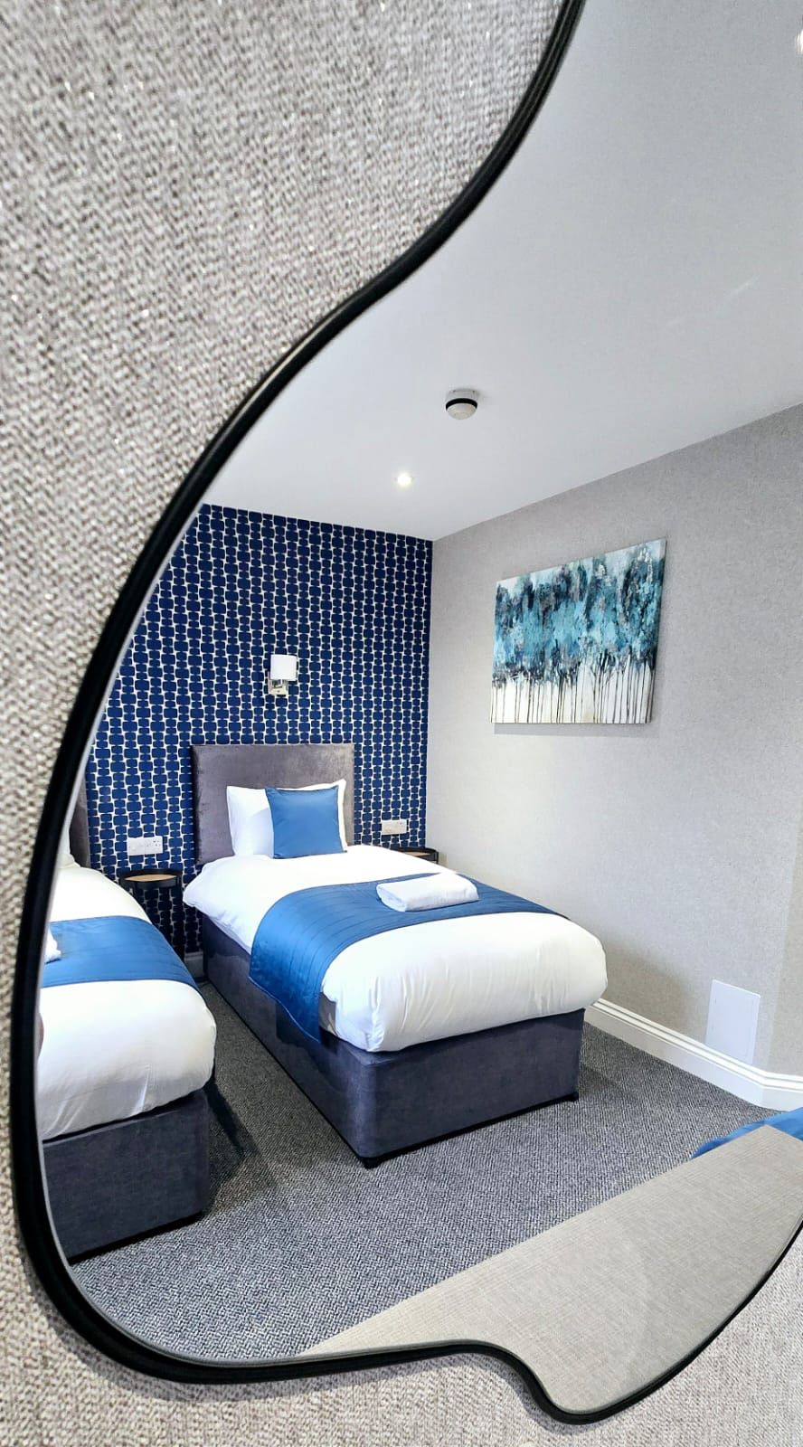 Mirror reflecting a hotel room with two twin beds. Blue and white bedding, blue patterned wall, and gray carpet.