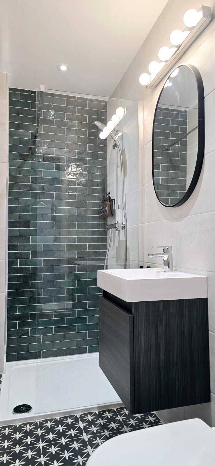A modern bathroom with a dark blue tile shower, a vanity, an oval mirror, and a patterned floor.