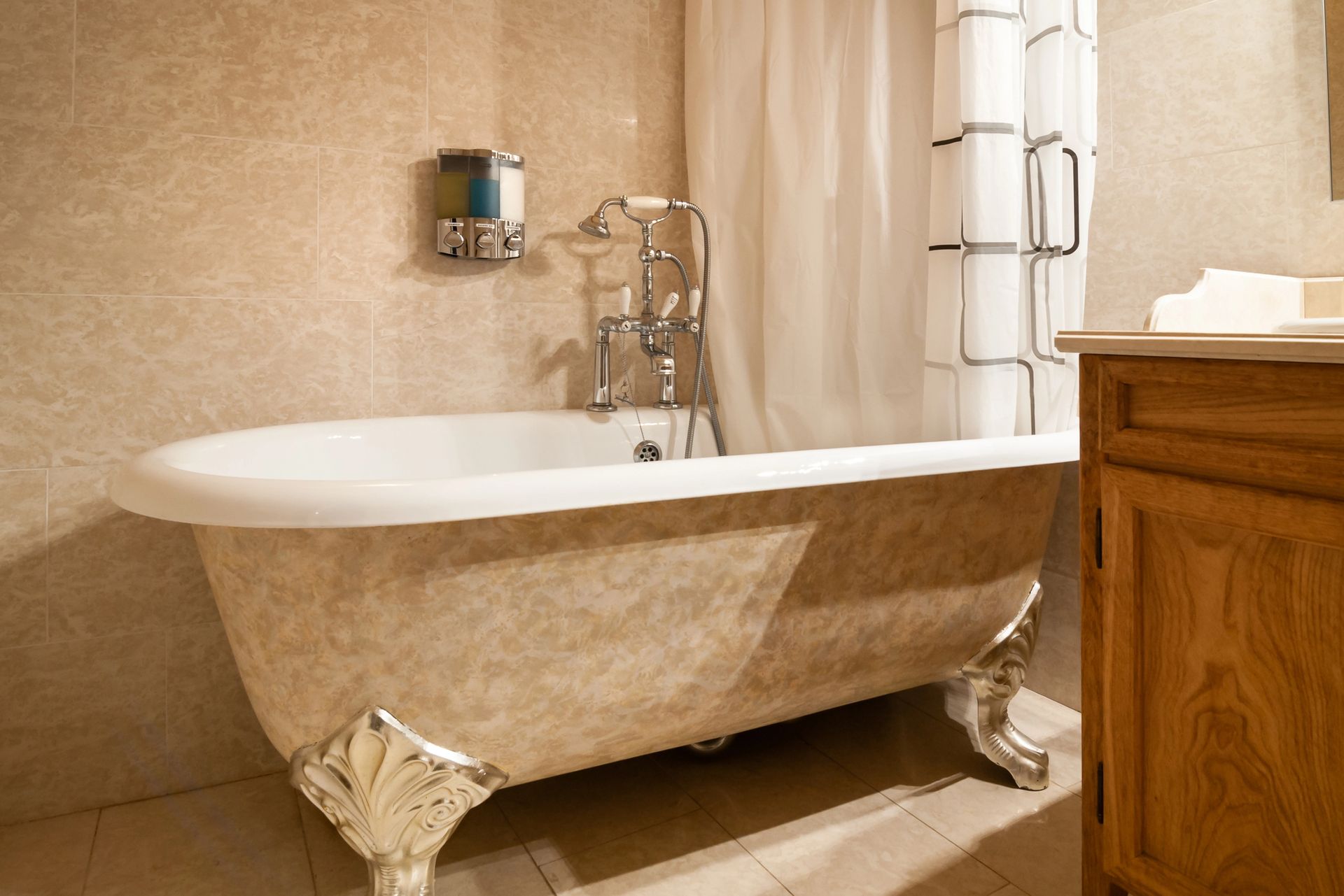 Vasca da bagno con piedini a zampa di leone e gambe decorate. Pareti piastrellate beige, tenda da doccia, mobiletto in legno e rubinetto cromato.