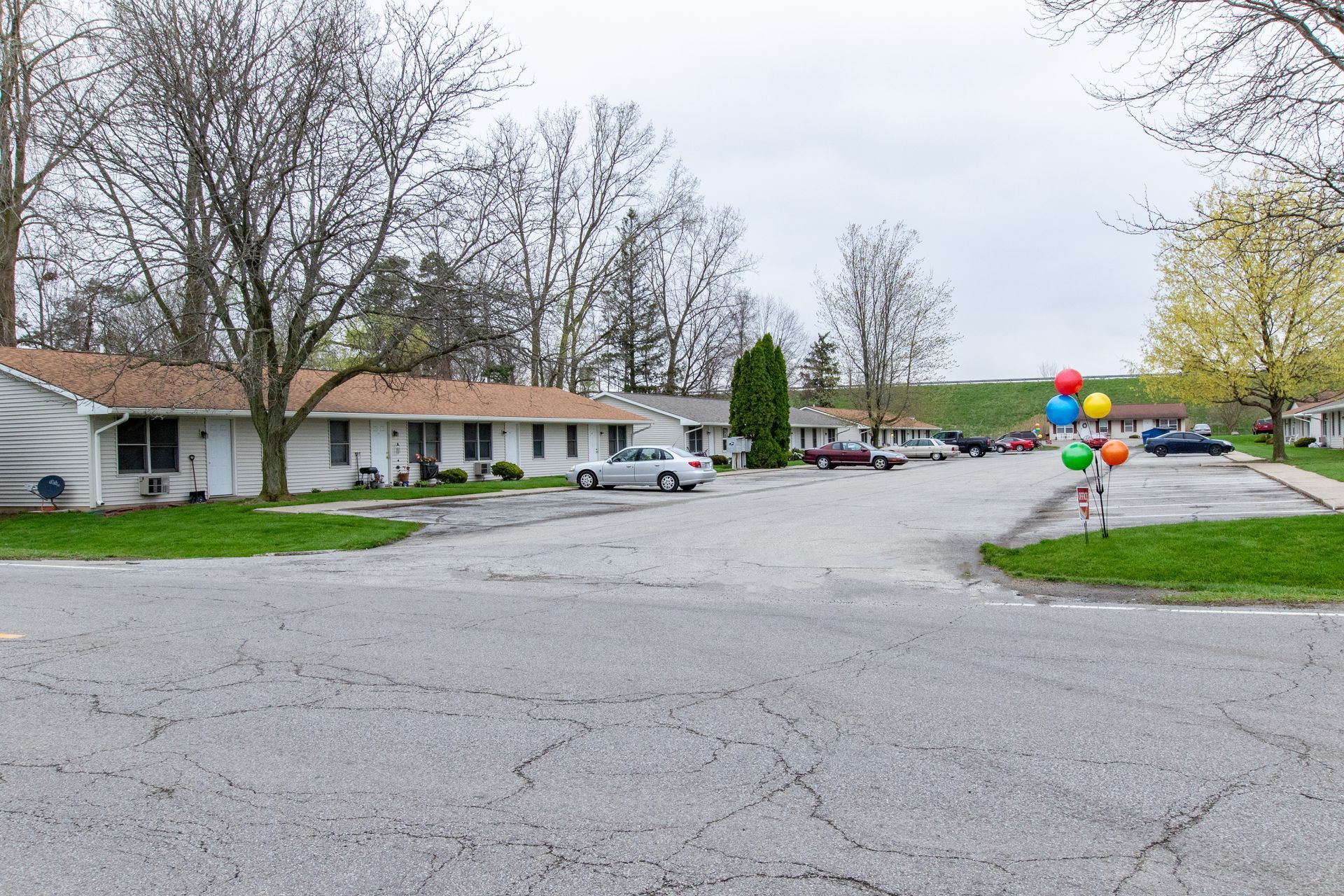 Apartment complex with a cracked asphalt road, cars, and colorful balloons. Overcast sky.