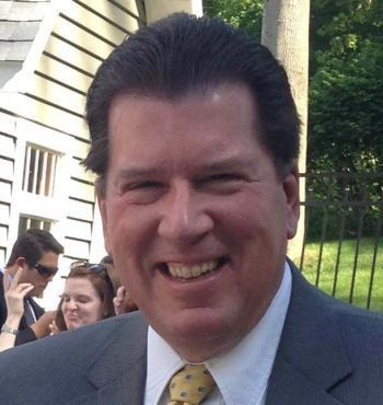 Smiling man in a gray suit and yellow tie outdoors, with people and a house in the background