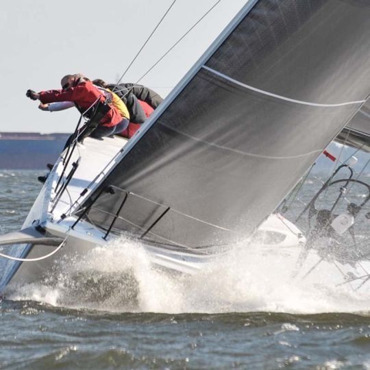 Two sailors racing a sailboat fast across choppy water, sail heeled sharply and spray splashing.
