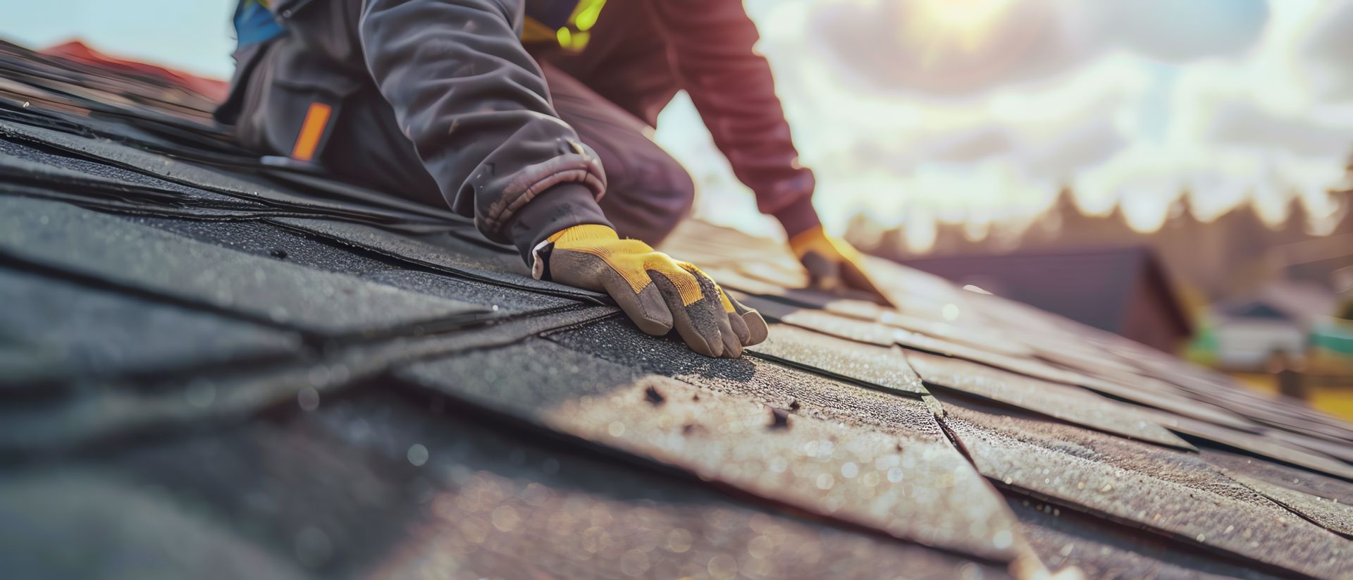 A person wearing work gloves installs dark shingles on a sloped roof under a bright, sunny sky.