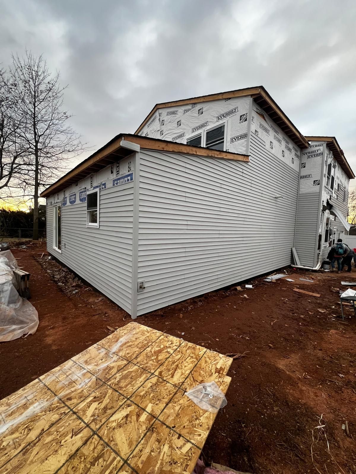 A house addition under construction with light gray horizontal siding, exposed framing, and red dirt ground.