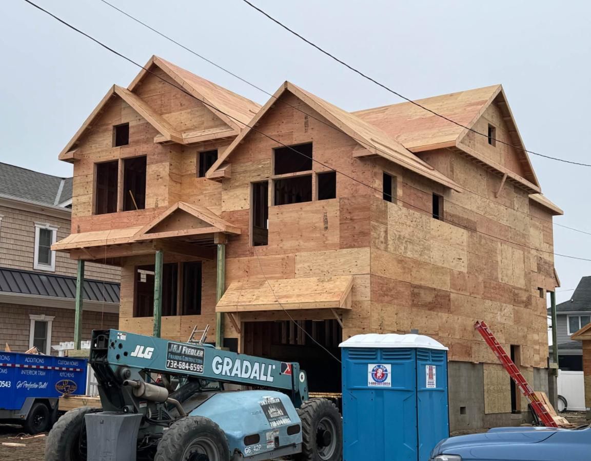 A two-story wood-framed house under construction with a blue Gradall telehandler and a portable toilet in the foreground.
