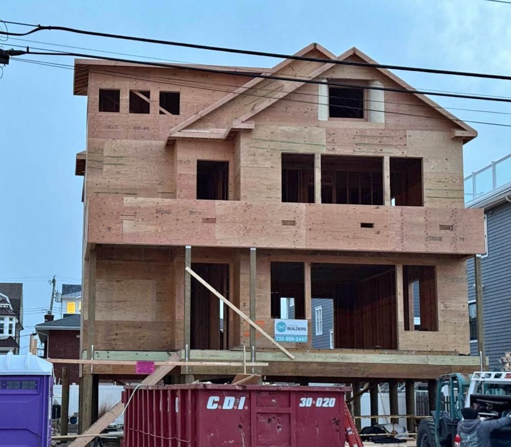 A three-story house under construction with exposed wooden framing, sheathing, and a large red dumpster in the foreground.