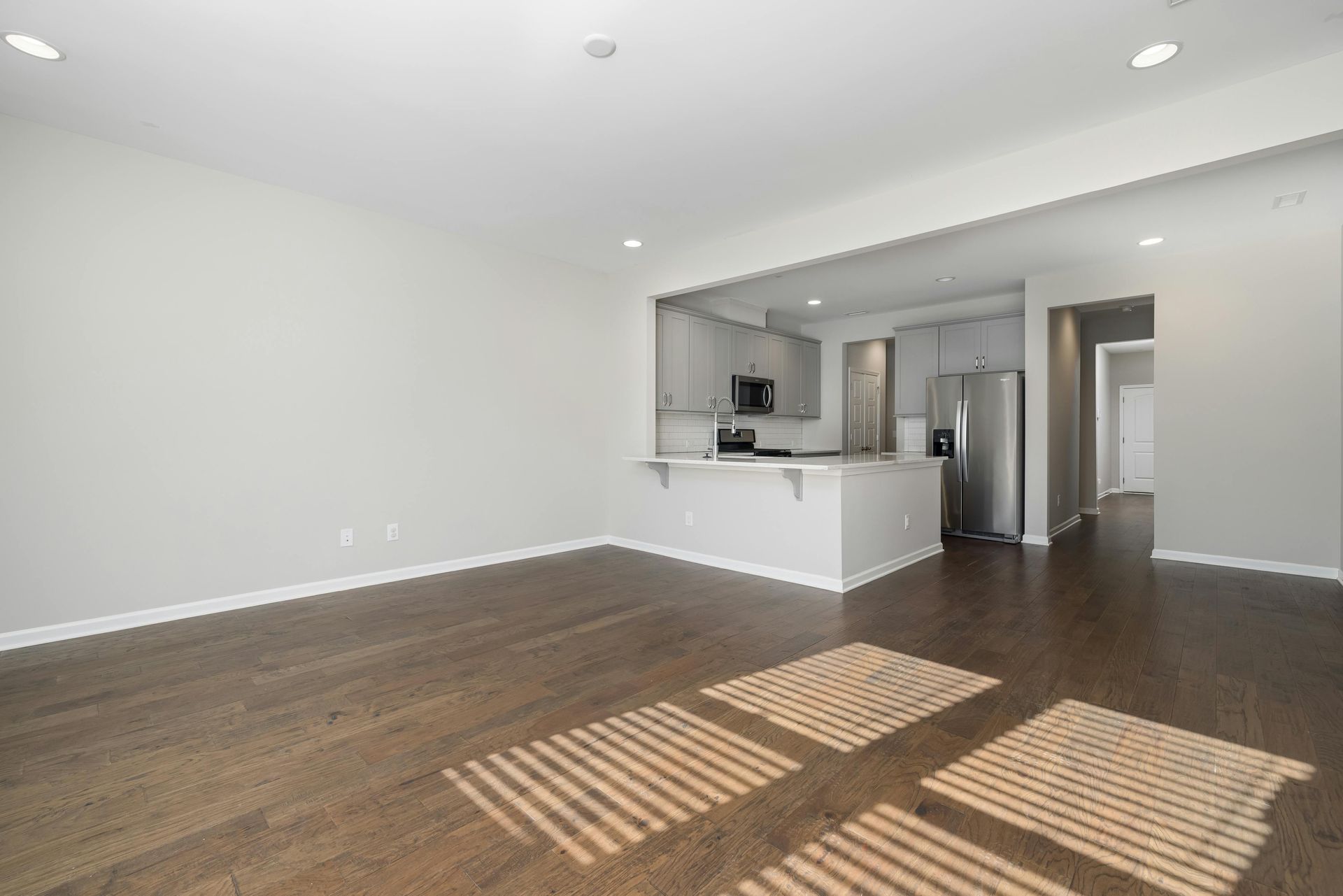 Empty modern living room with hardwood floors, white walls, and open kitchen with stainless steel appliances.