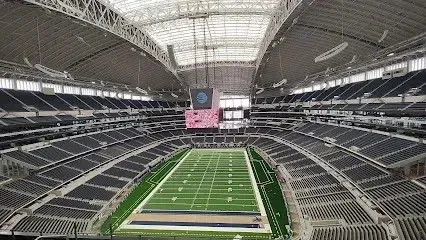 Burleson Electrician -
Interior view of a massive, modern football stadium with a retractable roof, tiered dark seating, and a large central video scoreboard above the green turf field.