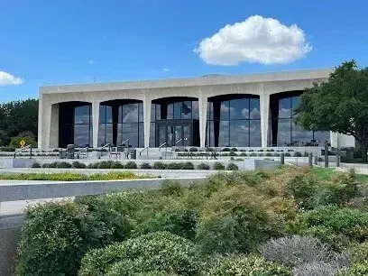 Burleson Electrician - Modern building with large glass windows framed by arches, set against a bright blue sky with fluffy clouds. The foreground features lush greenery.
