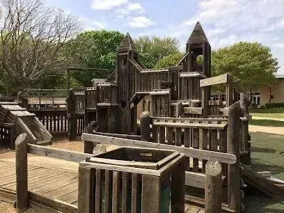 Burleson Electrician -Wooden playground structure with towers and bridges under a partly cloudy sky. Surrounded by trees and open grassy area, evoking an adventurous feel.
