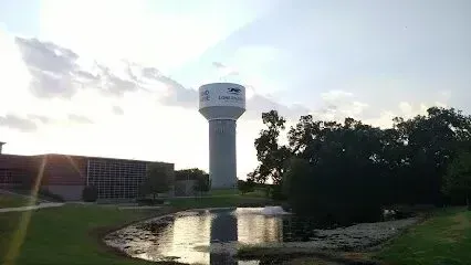 Burleson Electrician -A tall water tower with visible text stands over a grassy area with a pond reflecting the sky, near a modern building under a cloudy bright sky.