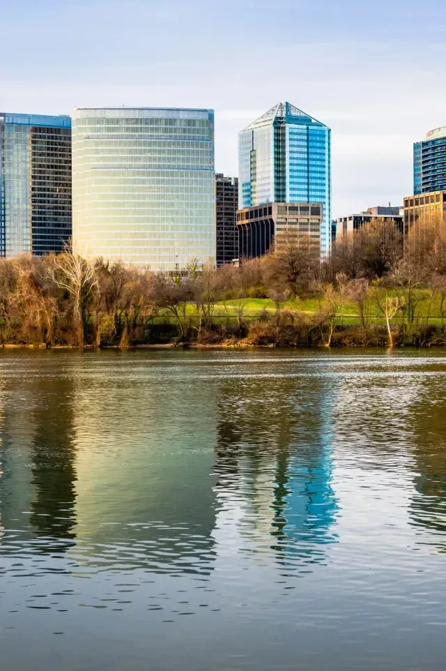 Burleson Electrician-Modern city skyline with tall glass skyscrapers reflected in the calm water of a river bordered by trees under a bright sky.