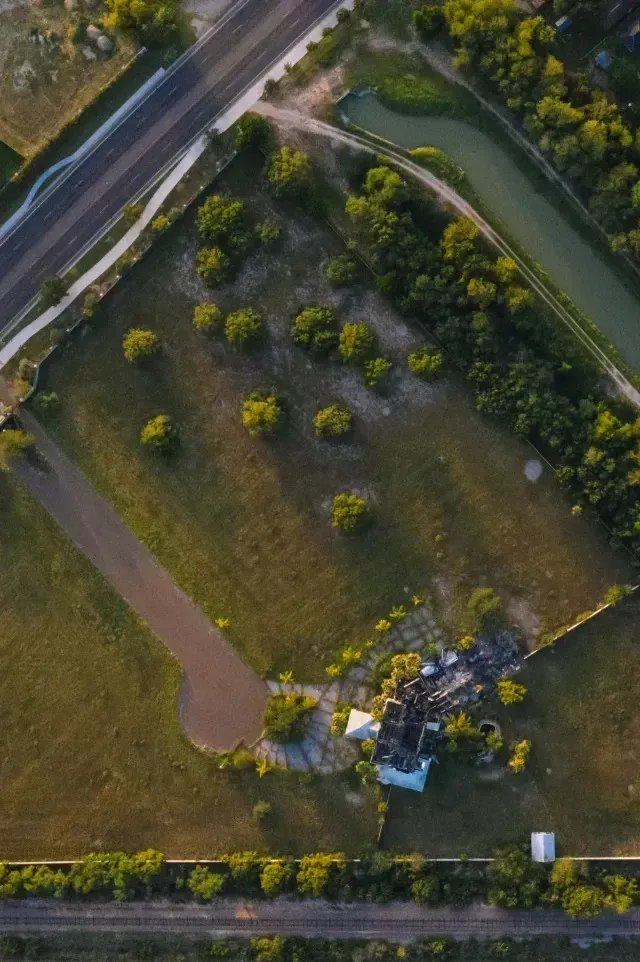 Burleson Electrician-Aerial view shows a grassy field with scattered trees, a partially destroyed structure, a curved dirt path, a highway, a canal, and parallel railroad tracks below.
