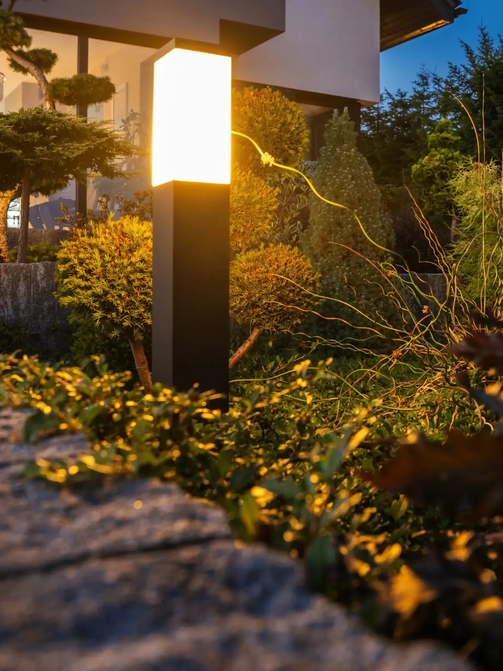Burleson Electrician - Modern black outdoor bollard light illuminates lush green and yellow foliage beside a stone retaining wall at dusk.