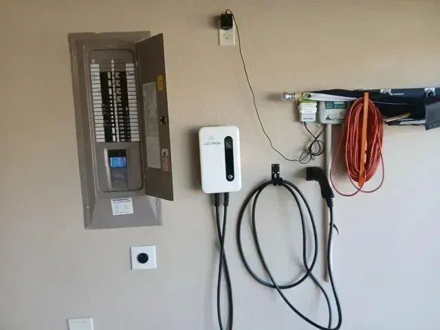 Burleson Electrician - A wall-mounted electrical panel beside an EV charger with charging cables, an electrical outlet, and tools hanging nearby.