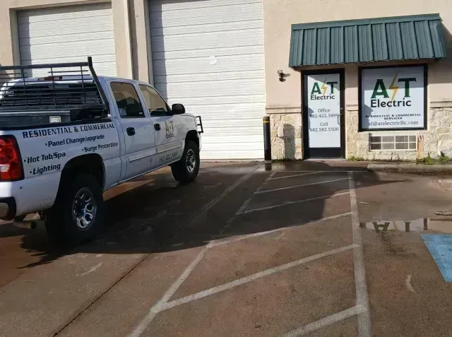 Burleson electrician - A white truck with a company logo parks outside A&T Electric's office, featuring a large door and signage for electrical services.