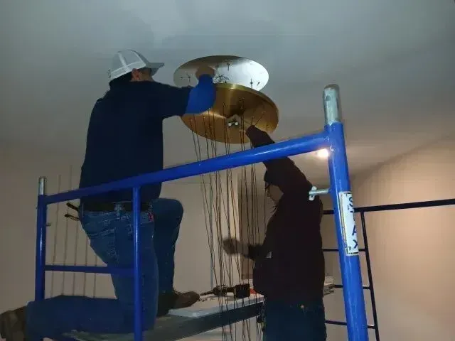 Burleson electrician - Two workers on blue scaffolding install a large, multi-tiered brass and white ceiling light fixture indoors.