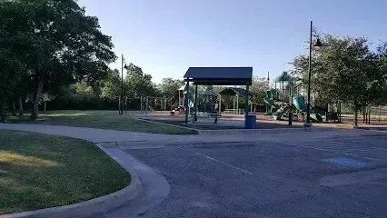 Burleson Electrician - Empty park playground area with a shaded structure, green play equipment, and a paved parking lot in the foreground under a bright sky.