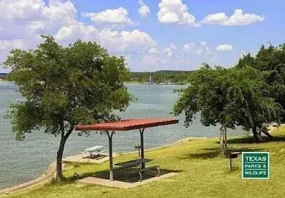 Burleson Electrician -A lakeside picnic area with a red-roofed shelter and bench in a grassy park, shaded by trees. A boat floats on the water under a blue sky.
