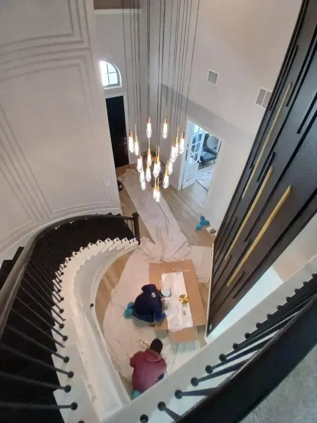 Burleson electrician - An overhead view of a spiral staircase, featuring two workers painting on a tarp below a modern chandelier in a well-lit entryway.