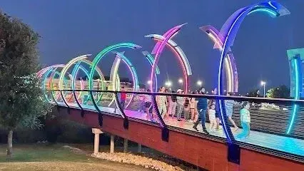 Burleson Electrician - Pedestrians walk across a bridge illuminated by colorful, glowing arched light fixtures against a dark blue twilight sky.