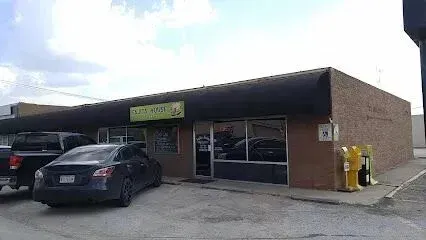 Burleson Electrician -A small, single-story brick store with a black awning and a green sign. Two cars are parked in front, with yellow newspaper dispensers nearby.