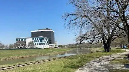Burleson Electrician - Clear blue sky over a river reflecting tall, dark corporate buildings across a grassy bank with a paved walking path and large, mostly bare trees in the foreground.
