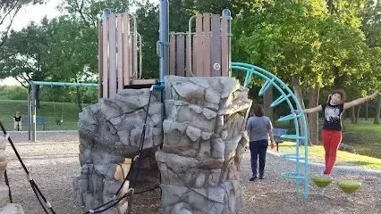 Burleson Electrician - Playground with rock-climbing structure, slide, and swings among trees. A child balances on stepping stones nearby, conveying joy and playfulness.
