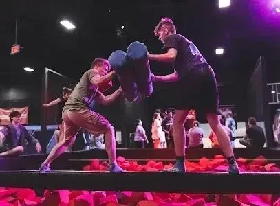 Burleson Electrician - Two people engage in a padded jousting match on a beam above a foam pit in a lively indoor play area. Onlookers and colorful lighting set an energetic tone.