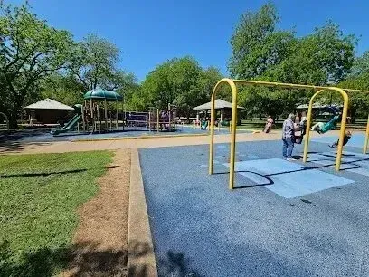 Burleson Electrician - Playground scene with swings and climbing equipment on a sunny day. Children play on blue rubber flooring, surrounded by trees and picnic areas.