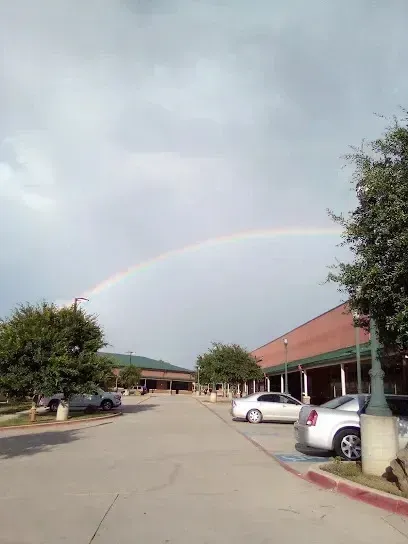 Burleson Electrician - A faint rainbow arches over a paved parking lot between two commercial buildings, with parked cars and green trees visible under a cloudy sky.