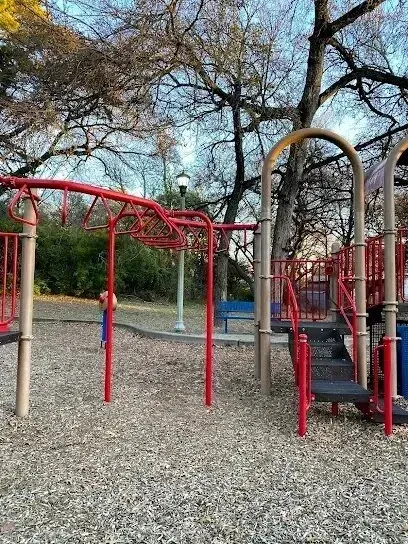 Burleson Electrician - Playground with red and tan climbing structures and monkey bars, set on wood chips. Leafless trees surround, under a clear blue sky.