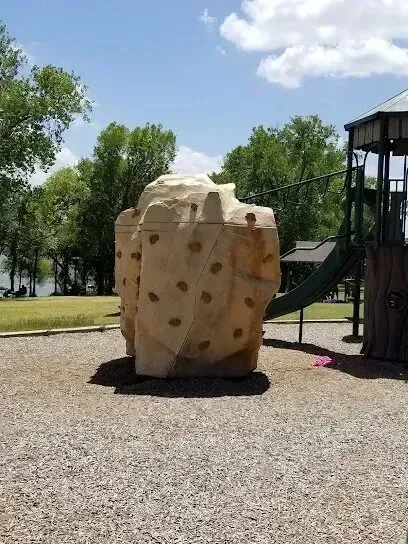 Burleson Electrician - A beige, rock-shaped climbing structure sits on wood chip ground at a sunny park near green trees and a playground slide.