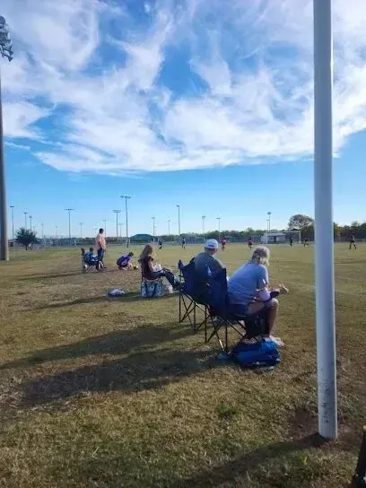 Burleson Electrician - Spectators sit in folding chairs on a grassy sports field under a bright blue sky with wispy white clouds, watching an event in the distance.