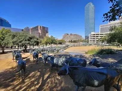 Burleson Electrician -Bronze cattle sculptures in a city park under a clear blue sky, surrounded by modern skyscrapers and trees, evoke a sense of history amid urban life.