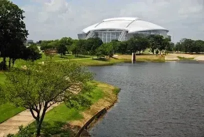 Burleson Electrician -A view across a dark body of water toward the large, white, domed AT&T Stadium surrounded by lush green parkland and trees under a bright, slightly cloudy sky.
