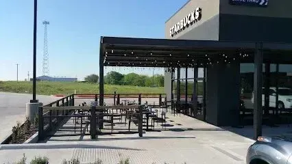 Burleson Electrician - Modern Starbucks location with an outdoor patio area featuring a dark pergola and seating, set against a backdrop of an open field and a distant cell tower under a clear blue sky.
