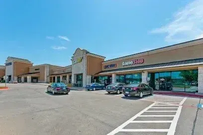 Burleson Electrician - Wide view of a sunlit shopping plaza with multiple storefronts, including GameStop. Cars are parked in front, and the sky is clear and blue.