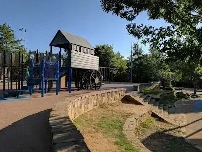 Burleson Electrician - A playground with a wooden structure resembling a train, blue slide, and steps. Surrounded by trees and a clear blue sky in the background.