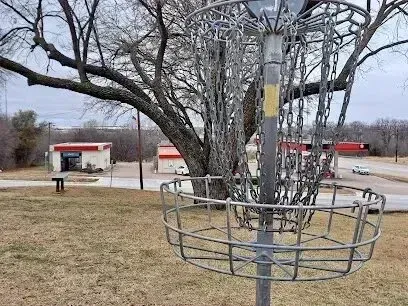 Burleson Electrician - A metal disc golf basket stands in the foreground against a backdrop of bare tree branches, brown grass, and commercial buildings near a road.