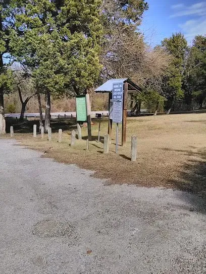 Burleson Electrician - Gravel path leads to wooden signs amid tall trees in a park. Sunlit grass and road in background create a serene and inviting atmosphere.