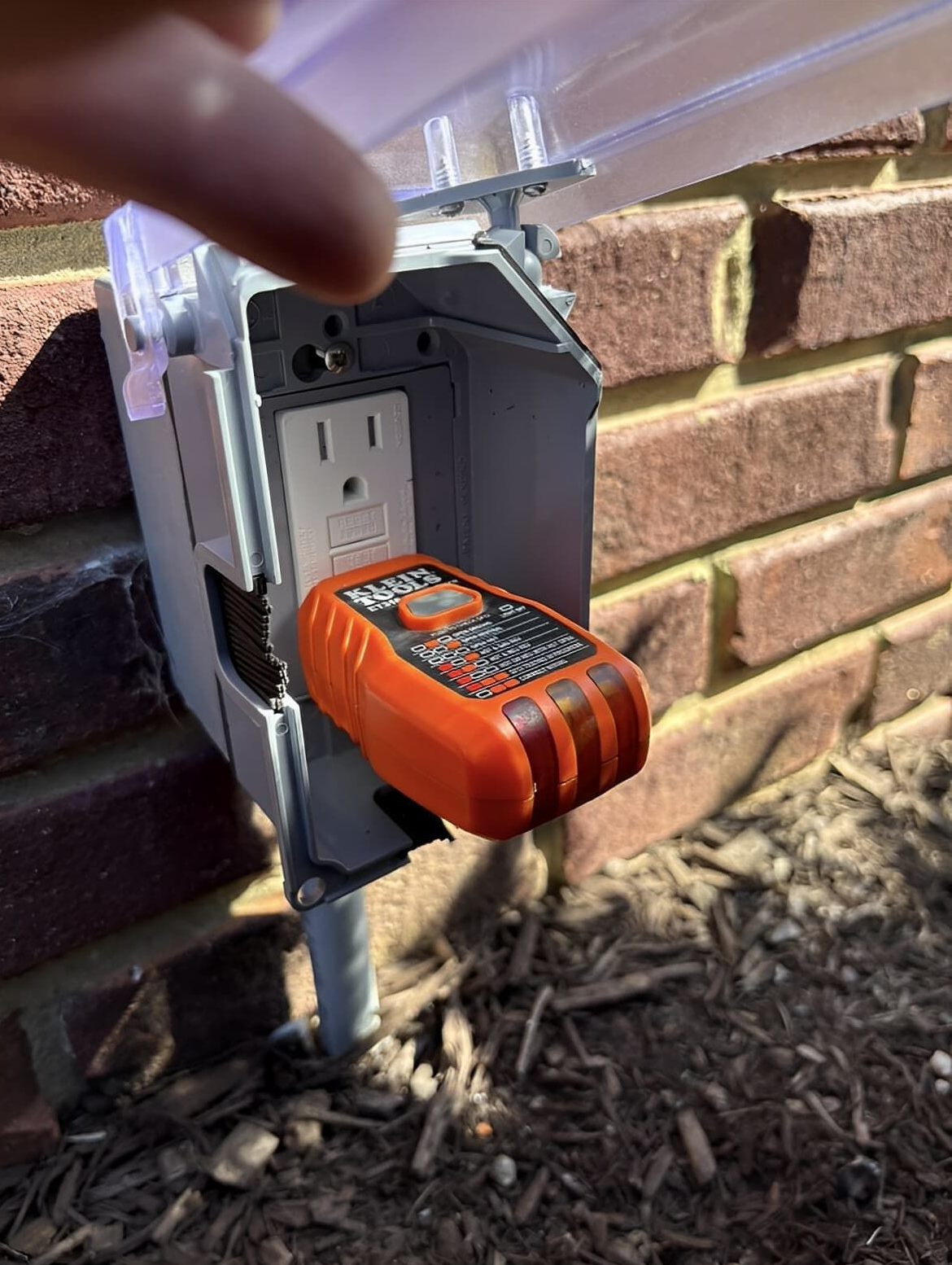 An electrical outlet tester inserted into an outdoor electrical outlet on a brick wall. A finger points at the outlet.