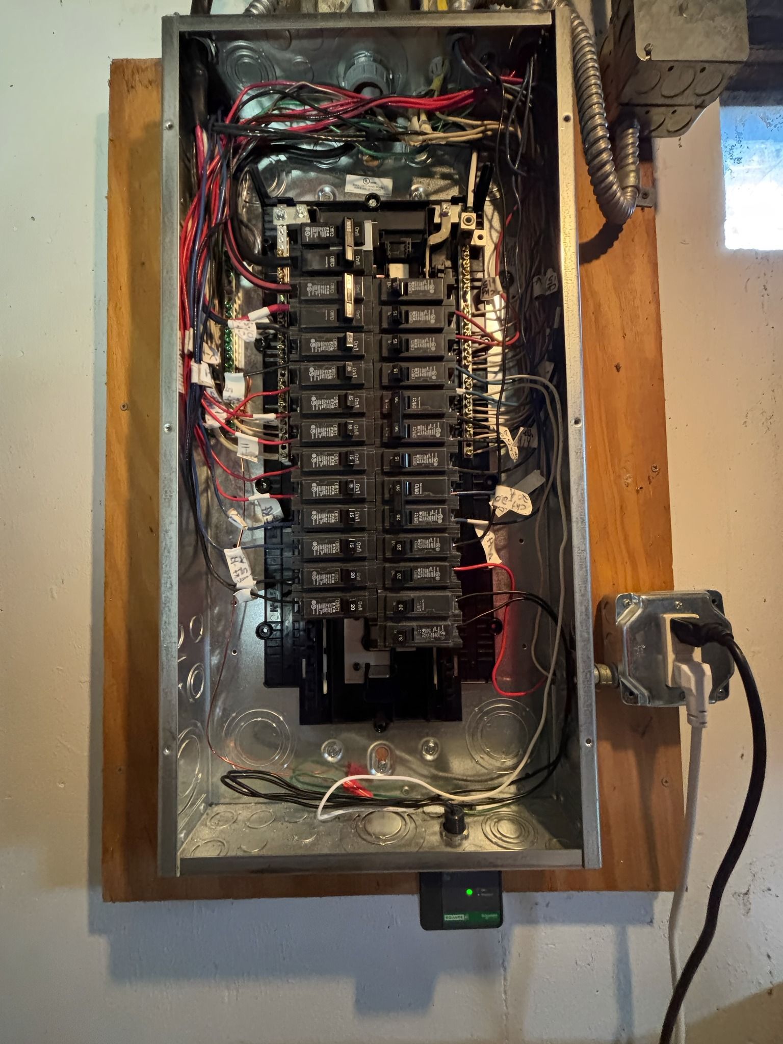 Electrical panel mounted on wood, containing circuit breakers and wiring. Black cord plugged into a nearby outlet.