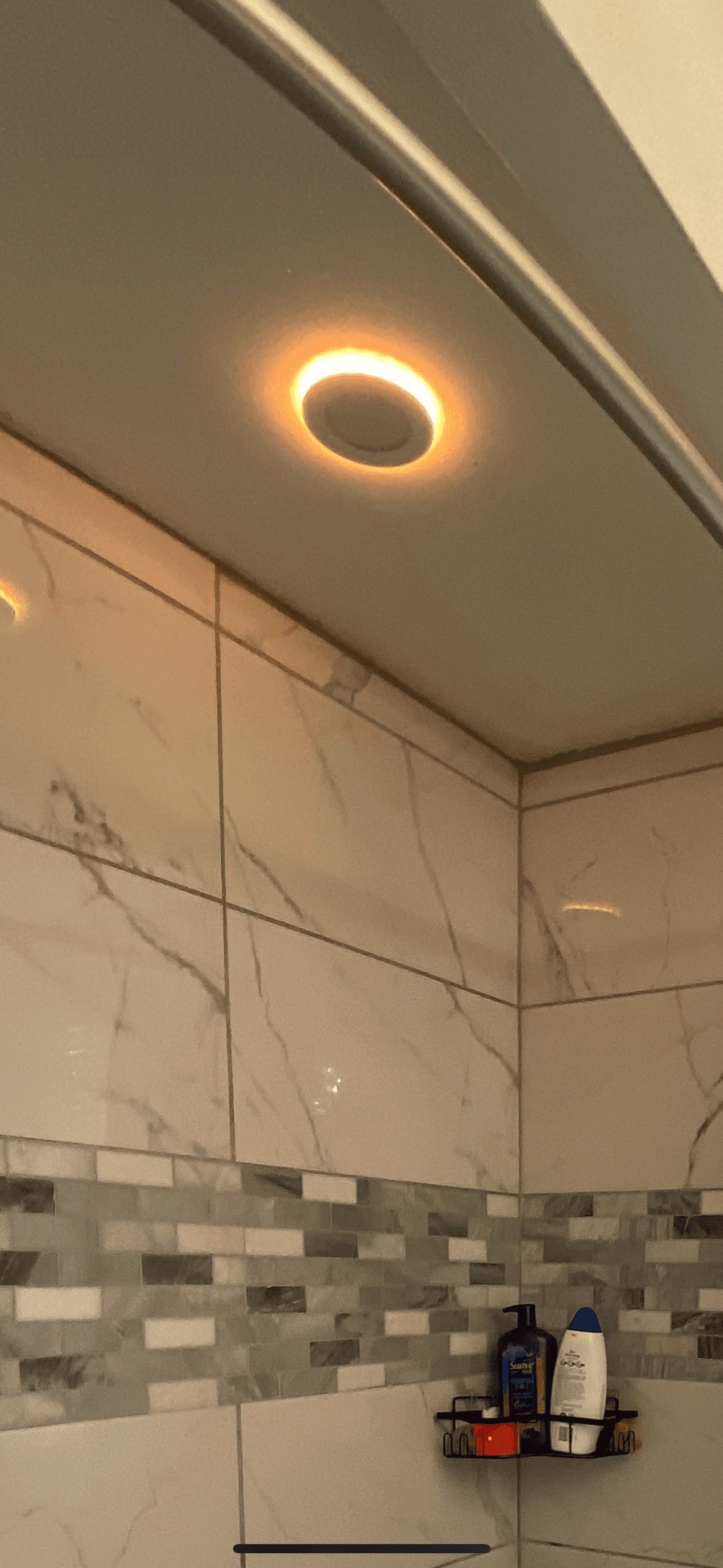 A bathroom with a lit ceiling light and marble tile. A shelf with toiletries is visible.