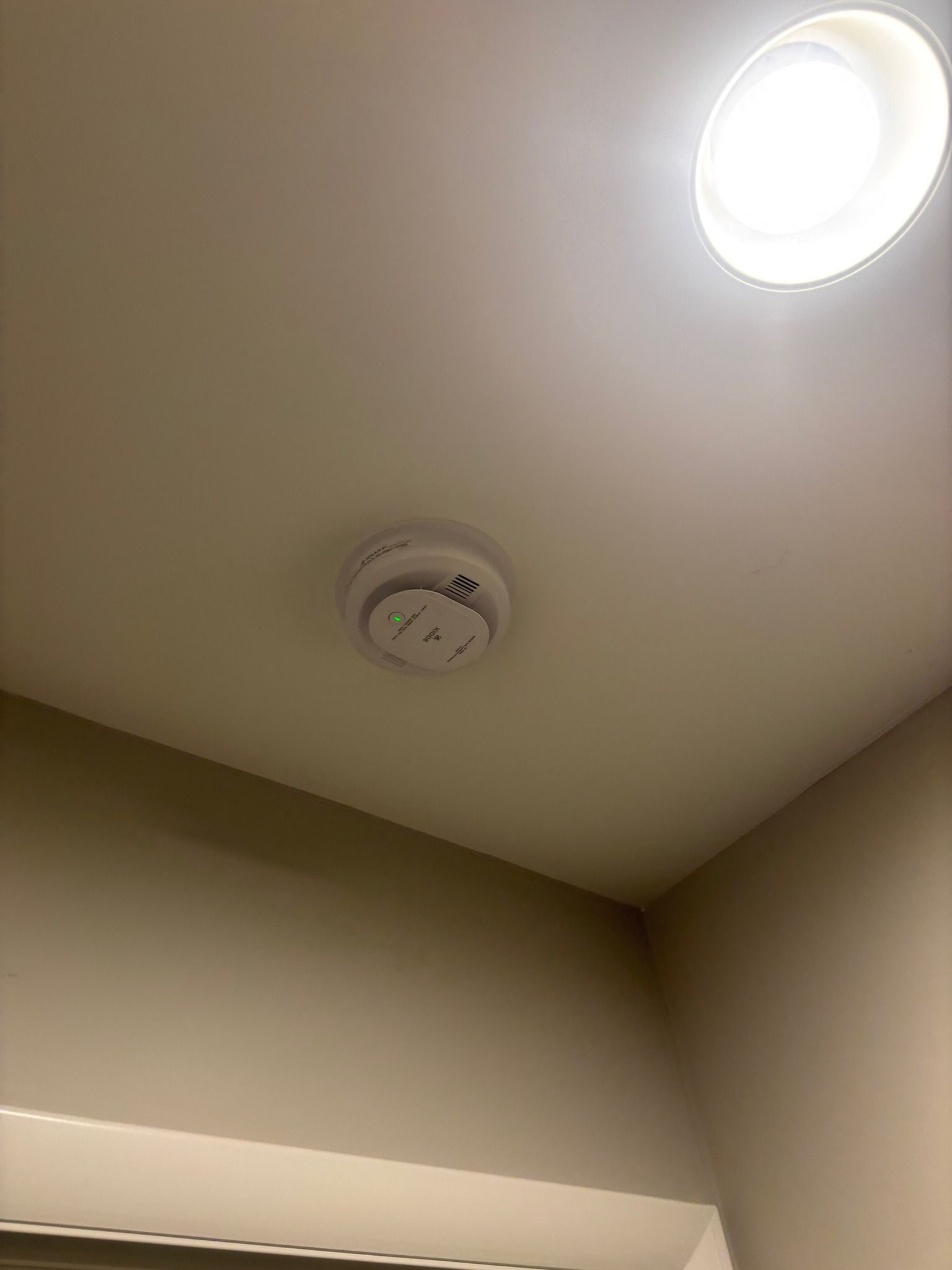 Smoke detector on a white ceiling with a circular light fixture in the corner of a room.
