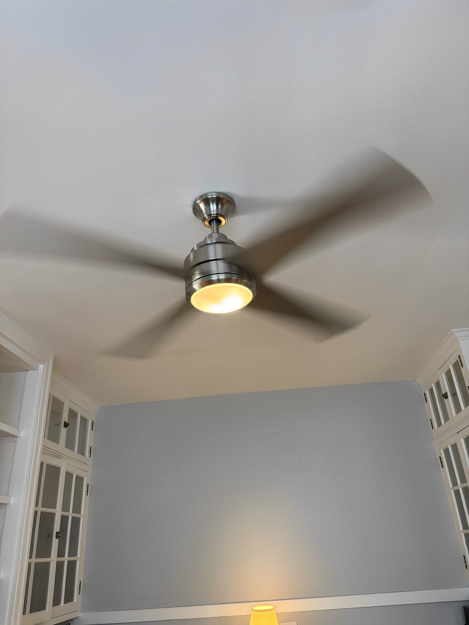 Ceiling fan with light on, casting shadows on a light blue wall, in a room with built-in shelves.