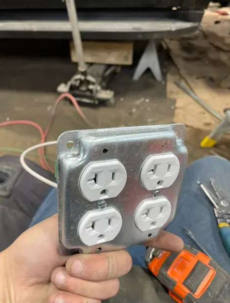 A hand holding a metal electrical box with four power outlets, wires attached.